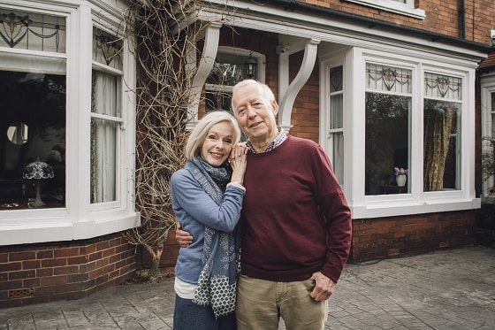 Senior couple are smiling for the camera while standing in front of their home.